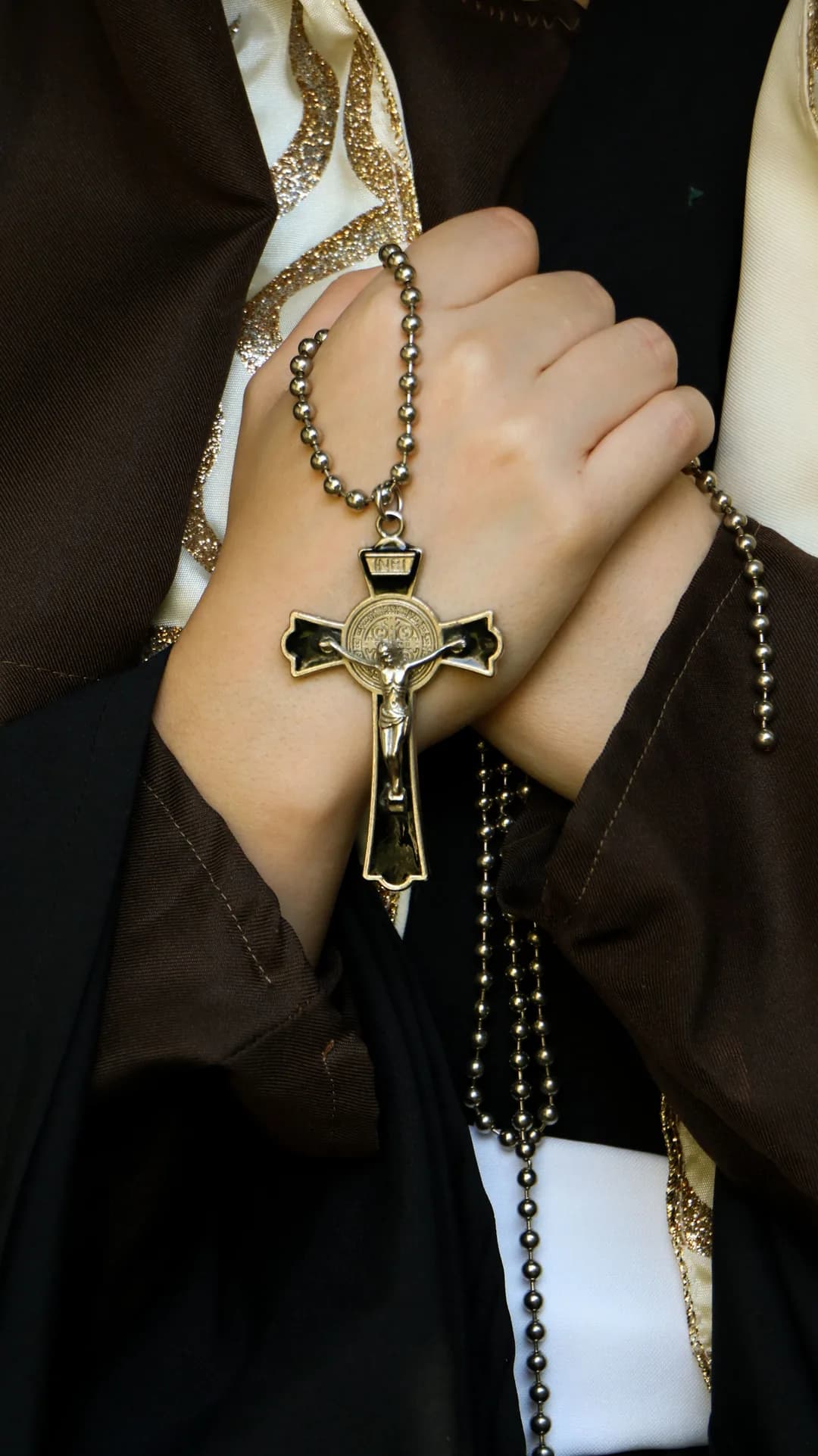 A pair of hands clasped in prayer, holding a Saint Benedict crucifix and rosary.