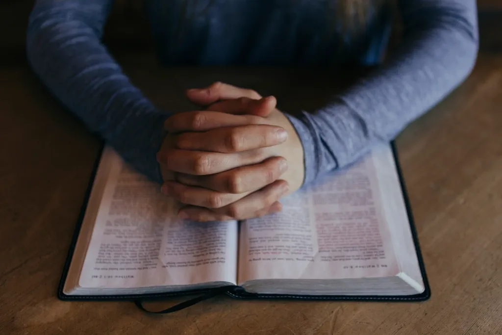 Hands joined in prayer resting on an open Bible.